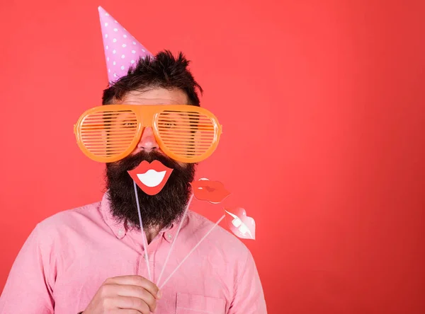 O tipo de chapéu de festa festeja, posando com adereços fotográficos. Hipster em óculos de sol gigantes comemorando. Homem com barba na cara alegre mantém lábios sorridentes em paus, fundo vermelho. Conceito de diversidade emocional — Fotografia de Stock