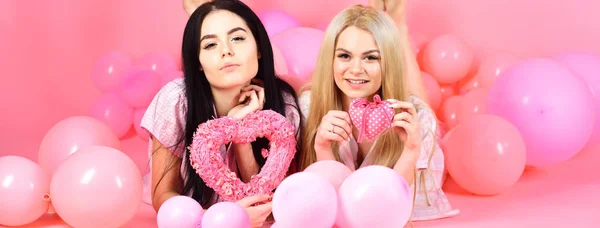 Brunett och blonda söta kvinnor i rosa pyjamas poserar på kamera med rosa hjärtan samtidigt som låg nära air ballonger på pyjamasparty över rosa bakgrund. — Stockfoto