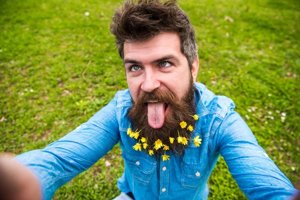 Mann mit Schöllkraut-Blüten im Bart macht Selfie-Foto. Hipster mit fröhlicher Fratze und Zunge auf dem Gesicht, der ein Selfie macht. Der Mensch genießt den Frühling, grünen Hintergrund, defokussiert. Heitere Stimmung — Stockfoto