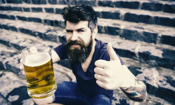 Piwo w warzywniaku. Człowiek z broda i wąsy posiada szkło z piwa i pokazuje kciuk w górę, schody kamienne podłoże. Piątek zrelaksować się koncepcja. — Zdjęcie stockowe