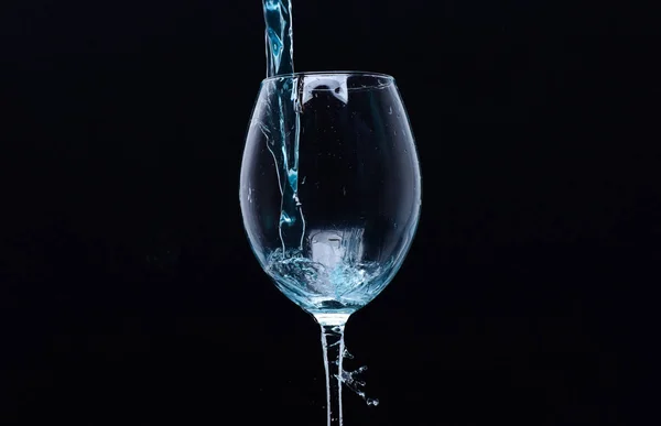Refreshing drink concept. Cocktail with blue liquid in glass. Glass with blue water pouring with liquid with splashes and drops. Wineglass filling with water with splashes on black background — Stock Photo, Image