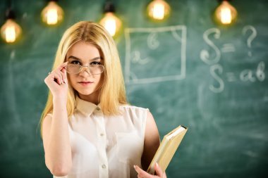 Öğrenci, öğretmen gözlük emin görünüyor, sınıfta, defocused arka plan, kara tahta hazır. Çekici öğretim görevlisi kavramı. Kadın kitap ile ders için hazırlar, izleyici olarak gazes