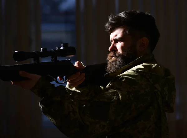 Man with beard wears camouflage clothing, dark interior background. Macho on suffering grimace face aiming at victim. Hunter, soldier with gun aiming before shooting. Shooter concept — Stock Photo, Image