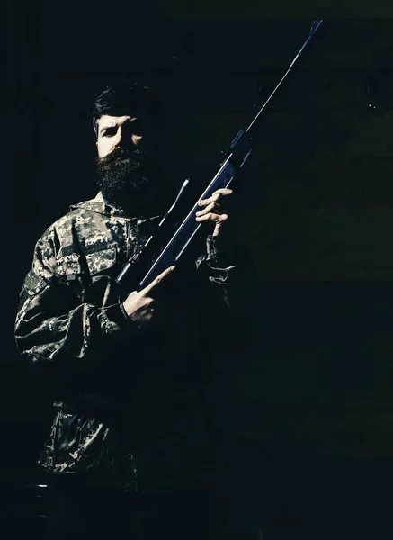 Macho en cara estricta en la casa de los guardabosques. Concepto de cazador. Hombre con barba lleva ropa de camuflaje, fondo interior de madera. Hunter, brutal hipster con arma lista para cazar —  Fotos de Stock