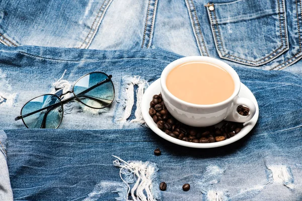 Caneca cheia de café com leite e óculos de sol em jeans, desfocada. Conceito de pausa para café. Xícara com café e feijão no prato no fundo denim. Cultura moderna de beber café — Fotografia de Stock
