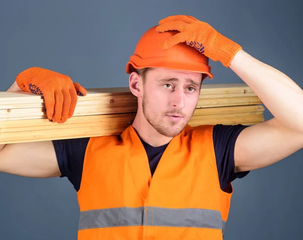 Ξυλουργός, ξυλουργός, ισχυρό οικοδόμος για σοβαρό πρόσωπο ασκεί ξύλινα δοκάρια στον ώμο. Έννοια ασφάλειας και προστασίας. Ο άνθρωπος στο κράνος, προστατευτικά γάντια κατέχει ξύλινο δοκάρι, σκληρό καπέλο και γκρι φόντο — Φωτογραφία Αρχείου