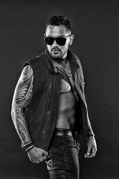 Fashion macho in trendy sunglasses. Bearded man with tattoo on chest and arms. Tattoo model with beard on unshaven face. Tattooed man with six pack and ab. Fashion style and trend, black and white