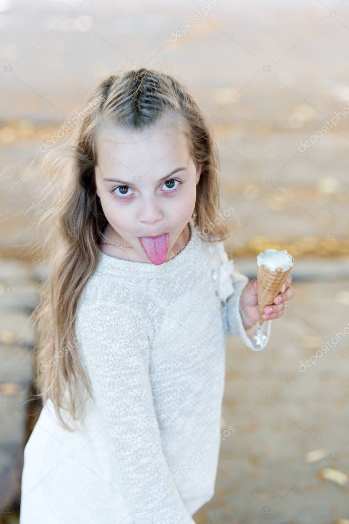 Girl sweet tooth on cheerful grimace with tongue eats ice cream, light background. Kid girl with ice cream cone in hand. Summer treats concept. Sweet tooth child with white ice cream in waffle cone