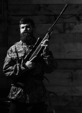 Avcı, acımasız hipster silahla avlamaya hazır. Maço gamekeepers evinde sıkı yüzünde. Avcı kavramı. Sakallı kamuflaj elbiseler, ahşap iç arka plan giyer