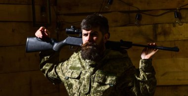 Erkeklik kavramı. Avcı, acımasız hipster silah avlamaya hazır ile sıkı yüzünde. Dostum, hakem sakallı kamuflaj elbiseler giyer, tüfek omuzlarında, ahşap iç arka plan taşır