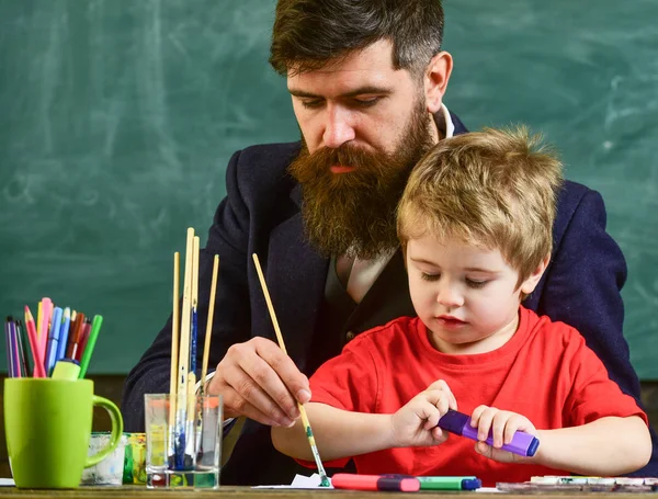 Teacher with beard, father and little son in classroom while drawing, creating, chalkboard on background. Talent and creativity concept. Child and teacher on busy faces painting, drawing