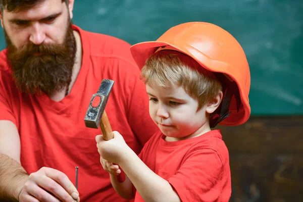 Rapaz, criança ocupada com capacete protector a aprender a usar martelo com o pai. Conceito de paternidade. Pai com barba ensinando o pequeno filho a usar ferramentas, martelando, quadro no fundo — Fotografia de Stock