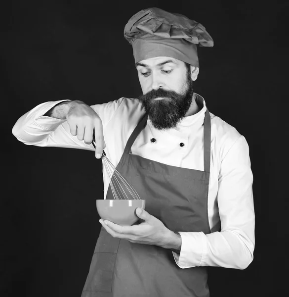 Cocinar con la cara curiosa en uniforme de color burdeos sostiene tazón, batir — Foto de Stock