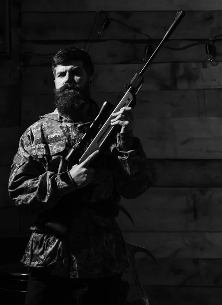 Hunter, um hipster brutal com uma arma pronta para caçar. Macho em rosto estrito na casa dos guarda-redes. Conceito de caçador. Homem com barba usa roupas de camuflagem, fundo interior de madeira — Fotografia de Stock