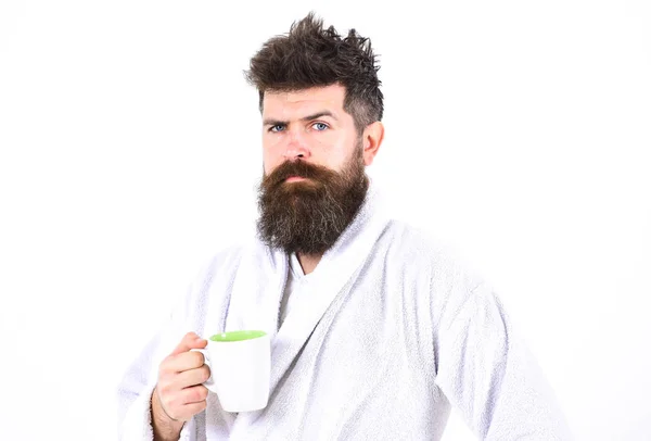 Hombre con barba y pelo desaliñado se levanta en albornoz, sostiene taza con té o café, fondo blanco. Concepto de rituales matutinos. Macho somnoliento, somnoliento con cara estricta bebe café en la mañana —  Fotos de Stock