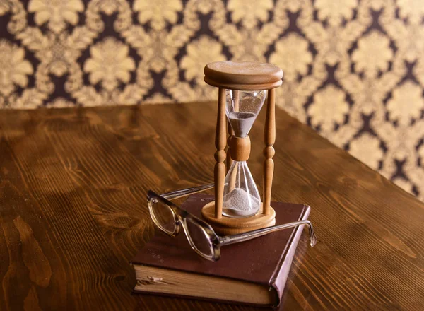 Пісочний годинник, стара книга та окуляри на дерев'яному столі, візерунок фону. Пісок падає всередину пісочного годинника. Концепція атрибутів мудрості. Годинник підраховує час біля старомодних окулярів — стокове фото
