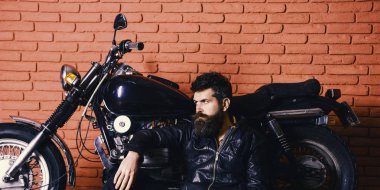 Vahşi motorcu kavramı. Yenilikçi, vahşi Motorcu deri ceketli dalgın yüzündeki motosiklet yakın yerde oturamaz. Sakallı, motorcu motorlu bisiklet garaj, tuğla duvar arka plan yakınındaki deri ceketli adam
