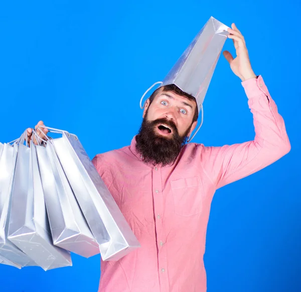 Uomo con barba e baffi porta borse della spesa, sfondo blu. Concetto di shopping. Guy shopping sulla stagione delle vendite con sconti. Hipster sul viso sorpreso con borsa sulla testa è dipendente shopaholic — Foto Stock