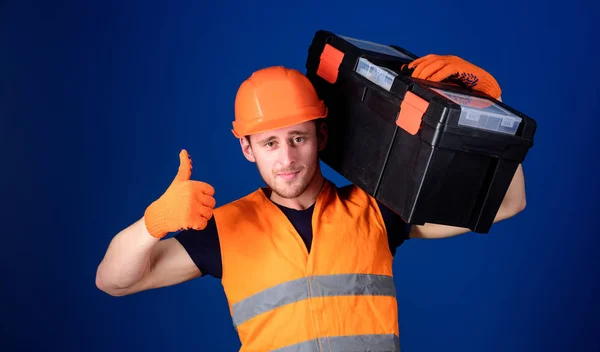 Homem de capacete, chapéu duro segura caixa de ferramentas e mostra gesto polegar para cima, fundo azul. Trabalhador, reparador, reparador, construtor no rosto confiante carrega caixa de ferramentas no ombro. Conceito de consulta de reparação — Fotografia de Stock