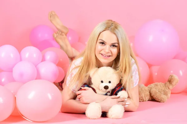 Girl in pajama, domestic clothes lay near air balloons, pink background. Blonde on smiling face relaxing with teddy bear toy. Woman cute celebrate birthday with balloons. Birthday girl concept