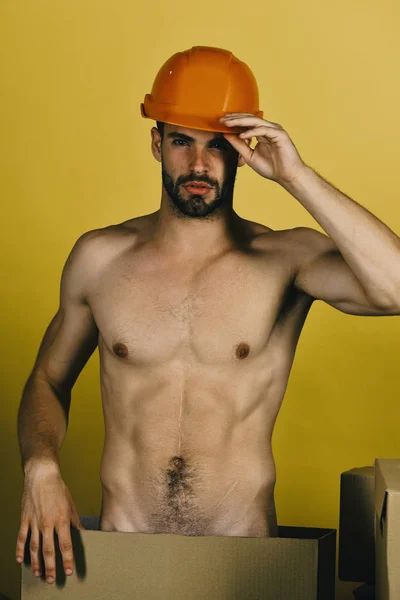 Hombre de pie desnudo entre cajas de cartón. Chico con torso sexy en sombrero duro sobre fondo amarillo — Foto de Stock