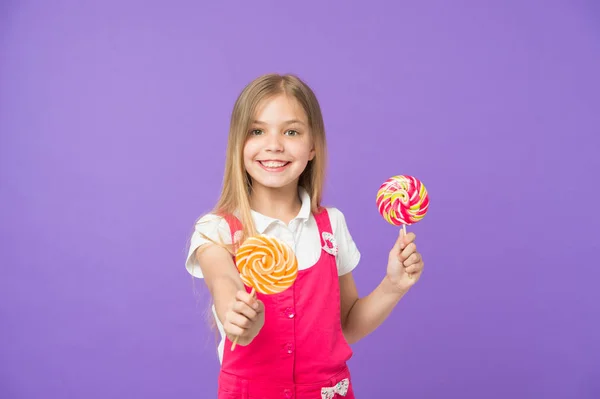 Comida y postre. Chica sonrisa con piruletas sobre fondo violeta. Niño feliz con caramelos de remolino sobre fondo púrpura. Pequeño niño sonriendo con caramelos en palos. Deliciosamente dulce. Dieta y dieta — Foto de Stock