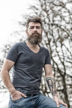 Ilık bahar günü parkta uzun sakal ayakta yorgun orta yaşlı adam. Dövmeli kolu koyu mavi T-shirt ve kentsel sahnede yürümek denim jeans giyen kaslı acımasız adam