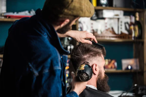 Hipster bearded client getting hairstyle. Barber with hairdryer drying and styling hair of client. Barber with hairdryer works on hairstyle for bearded man, barbershop background. Styling concept — Stock Photo, Image