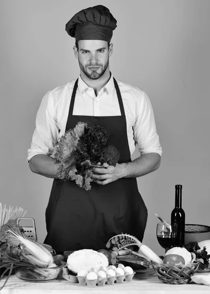 Mannen i kock hatt och förkläde med sallad. Cook fungerar i köket med grönsaker och verktyg. — Stockfoto