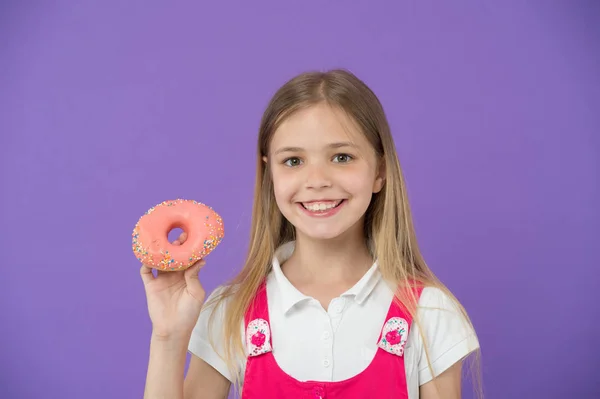 Do you want bite. Girl smiling face holds pink donut in hand, violet background. Kid smiling girl ready to bite donut. Snack concept. Child can not wait to eat sweet donut, copy space