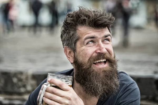 Hipster with long beard looks cheerful. Bearded hipster holds beer mug, drinks beer outdoor. Man with beard and mustache on happy face, urban background, defocused. Craft beer concept