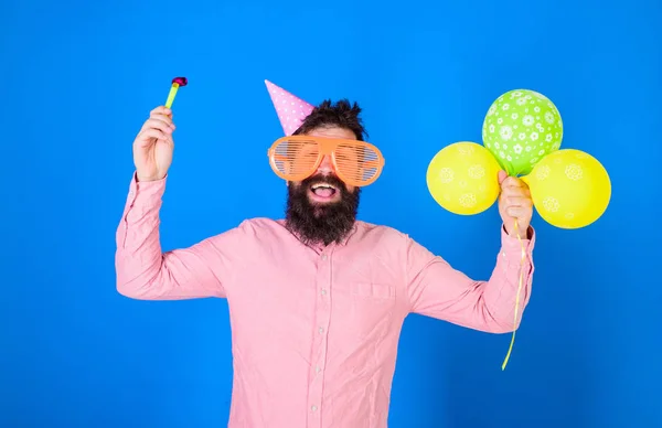 Hipster em óculos de sol gigantes comemorando aniversário. Homem com barba e bigode no rosto feliz detém chifre de festa, fundo azul. Conceito de celebração. Cara em chapéu de festa com balões de ar celebra — Fotografia de Stock