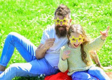 Father and daughter sits on grass at grassplot, green background. Superstar concept. Child and dad posing with star shaped eyeglases photo booth attribute at meadow. Family spend leisure outdoors clipart