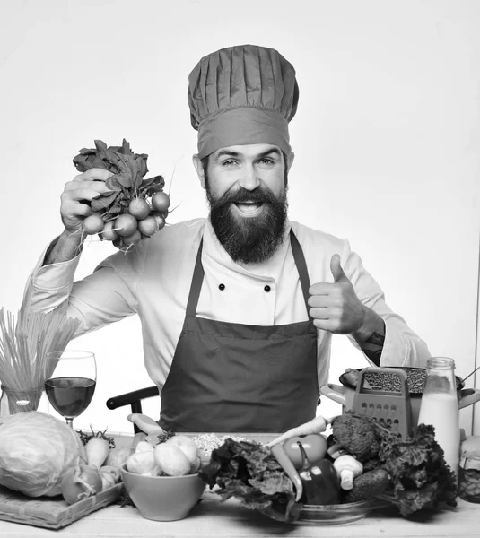 Cuire avec le visage heureux en uniforme assis à table avec des légumes et des ustensiles de cuisine. Homme à la barbe tient radis bouquet sur fond blanc — Photo