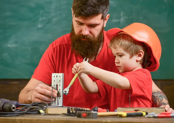 Boy, child busy in protective helmet makes by hand, repairing, does crafts with dad. Father with beard teaching little son to use tools in classroom, chalkboard on background. Mens work concept