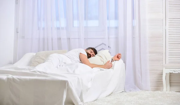 Guy on calm face sleeping on white sheets, pillow. Nap and siesta concept. Macho with beard sleeping, relaxing, having nap, rest. Man laying on bed, covered with blanket, white curtains on background — Stock Photo, Image