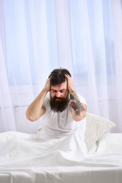 Hangover concept. Man in shirt sits on bed awake, white curtain on background. Guy on disappointed painful face waking up in morning. Macho with beard and mustache suffers from hangover