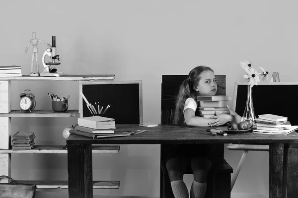 Κορίτσι που κάθεται στο ξύλινο γραφείο με πολύχρωμα χαρτικά, λουλούδια και φρούτα. Μαθήτρια με γκρινιάρης πρόσωπο κατέχει στοίβα βιβλίων — Φωτογραφία Αρχείου