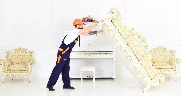 Loader moves sofa, couch. Delivery service concept. Man with beard, worker in overalls and helmet lifts up sofa, white background. Courier delivers furniture in case of move out, relocation