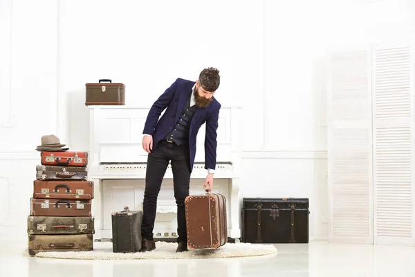 Macho elegante op strikte gezicht staat in de buurt van stapel van vintage koffer, klaar voor de vakantie. Man, reiziger met baard en snor met bagage, luxe witte interieur achtergrond. — Stockfoto
