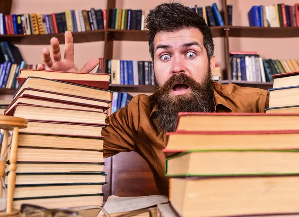 Homem no rosto chocado entre pilhas de livros, enquanto estudava na biblioteca, estantes de livros no fundo. Professor ou estudante com barba senta-se à mesa com óculos, desfocado. Conceito-limite — Fotografia de Stock