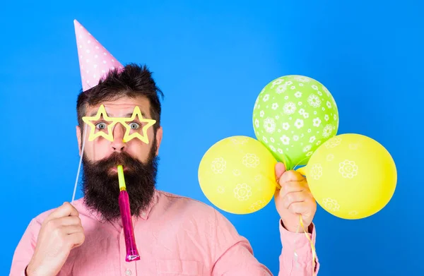 Mannen med skägg på förvånade ansikte innehar air ballonger, blå bakgrund. Killen i partiet hatt med semester attribut firar. Hipster i stjärna formad glasögon blåser in part horn. Överraskning-konceptet — Stockfoto
