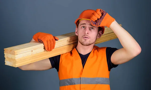 El hombre con guantes protectores sostiene visera de sombrero duro protector, fondo gris. Carpintero, carpintero, fuerte constructor en la cara seria lleva la viga de madera en el hombro. Concepto de seguridad y protección —  Fotos de Stock