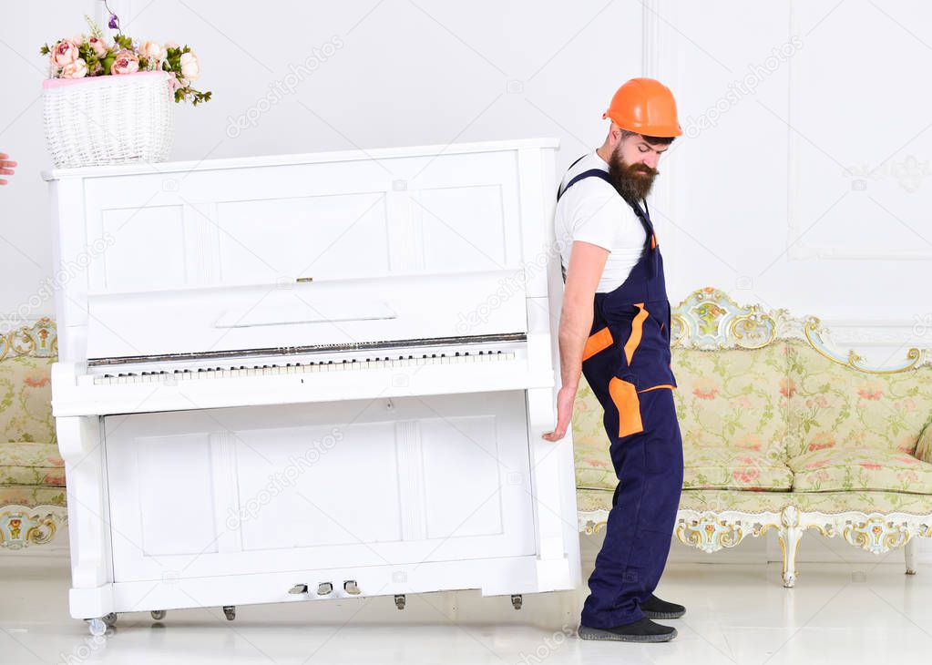 Loader moves piano instrument. Heavy loads concept. Man with beard worker in helmet and overalls lifts up, efforts to move piano, white background. Courier delivers furniture, move out, relocation