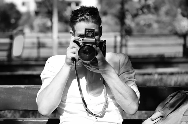 Человек держит фотокамеру на фоне зеленого парка. Молодой путешественник или фотограф сидит на скамейке возле рюкзака . — стоковое фото