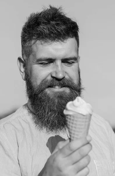 Man with long beard enjoy ice cream. Sweet tooth concept. Bearded man with ice cream cone. Man with beard and mustache on smiling face eats ice cream, blue sky background, defocused
