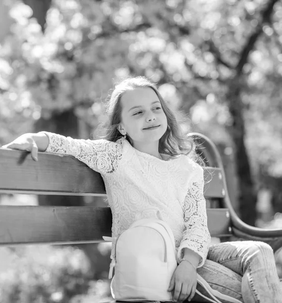 Doplňky koncept. Dívka na usmívající se tvář sedí na lavičce, strom sakura na pozadí, rozostření. Roztomilé dítě s batohem užívat slunný jarní den. Dívka relaxační při procházce v parku nedaleko třešňový květ — Stock fotografie
