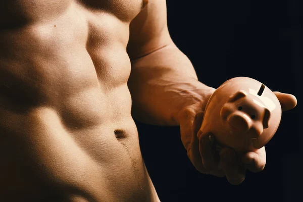 Guys abs and strong chest. Athletes body in close up, hand holding pink piggy bank. Male with naked torso isolated on black background. Sports, bodybuilding,