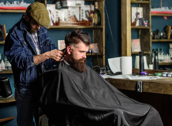 Barber with hair clipper works on haircut of bearded guy, retro barbershop background. Hipster hairstyle concept. Hipster client getting haircut. Barber with clipper trimming hair on nape of client