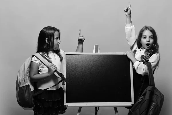 Schoolgirls next to chalkboard on pink background. Girls with surprised faces hold fingers up having idea. Kids wearing schoolbags lean on blackboard, copy space. — Stock Photo, Image
